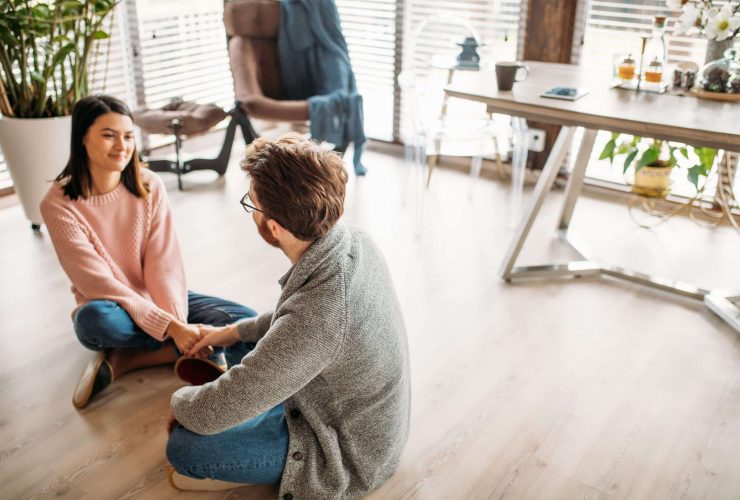 Man and woman sitting on the floor holding hands in conversation, representing supportive peer connection in recovery aftercare.
