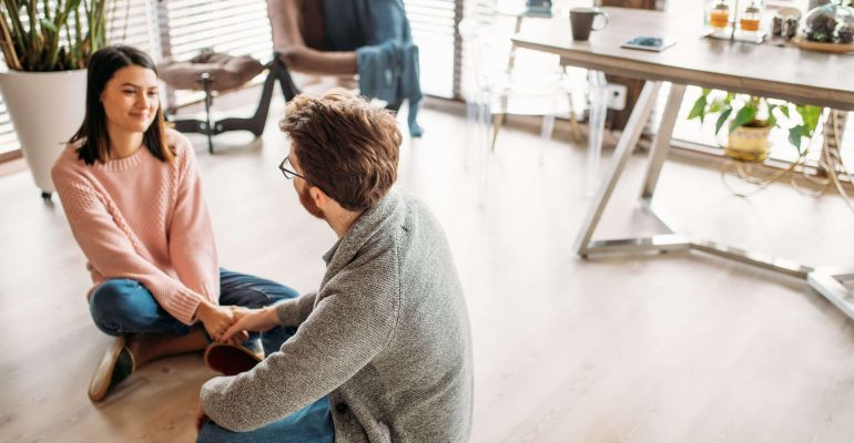 What Role Does Aftercare Play in the Success of Recovery Programs? 3 Man and woman sitting on the floor holding hands in conversation, representing supportive peer connection in recovery aftercare.