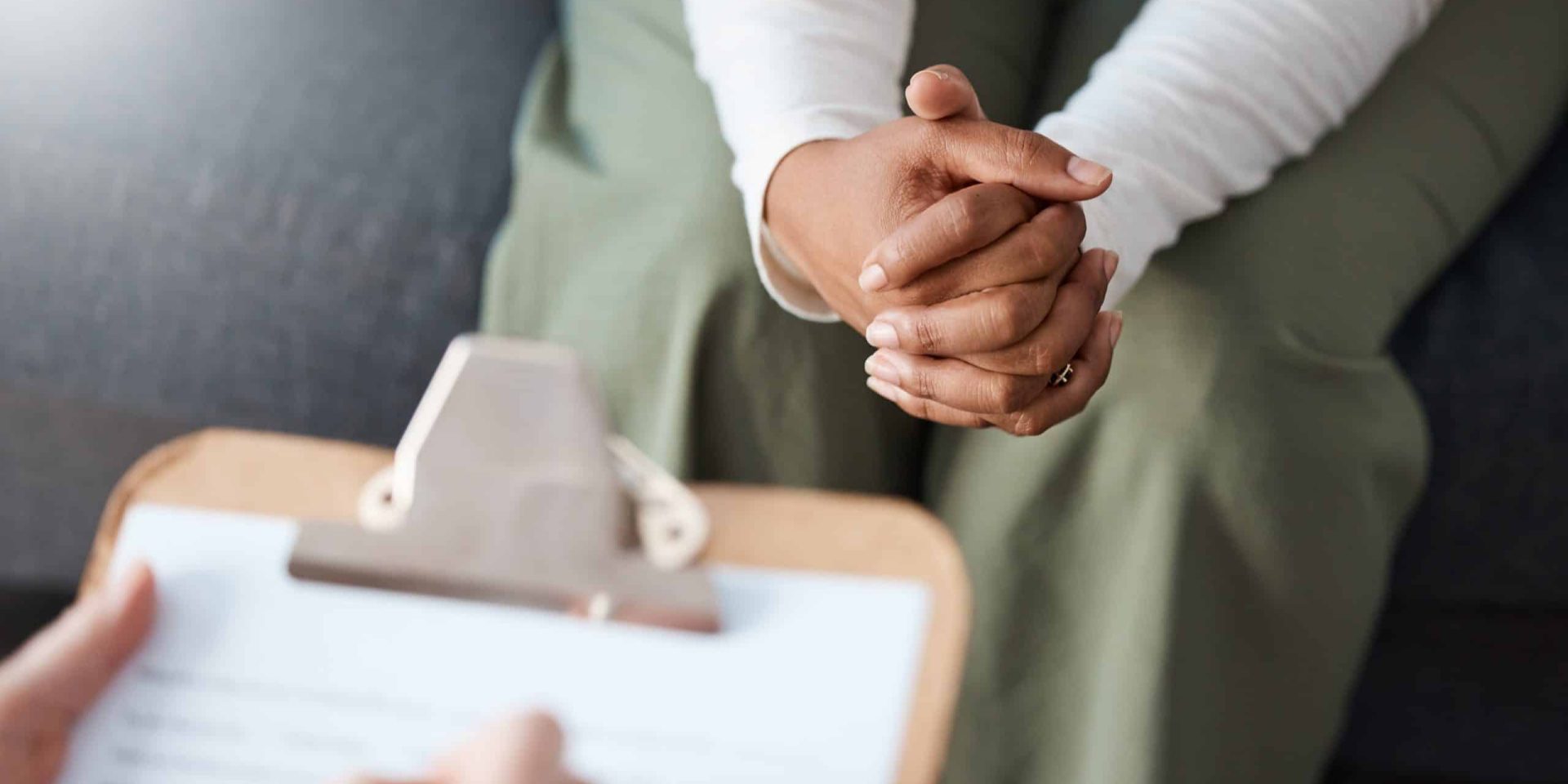 Client with clasped hands during a one-on-one counselling session, with a clinician's clipboard visible in the foreground